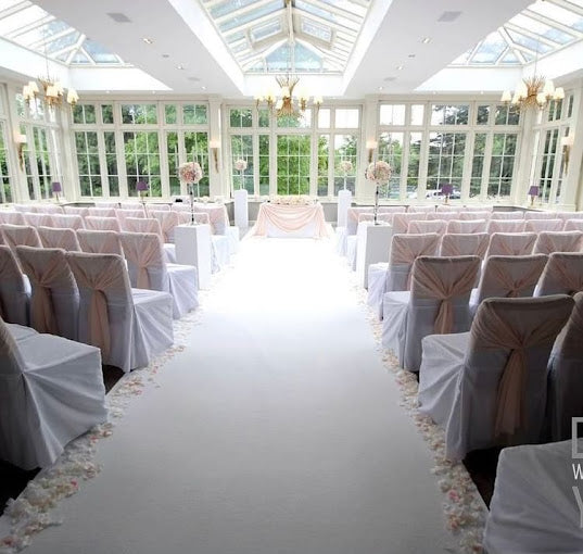Elegant wedding venue setup with white chairs and pink sashes, featuring a floral aisle and bright windows.